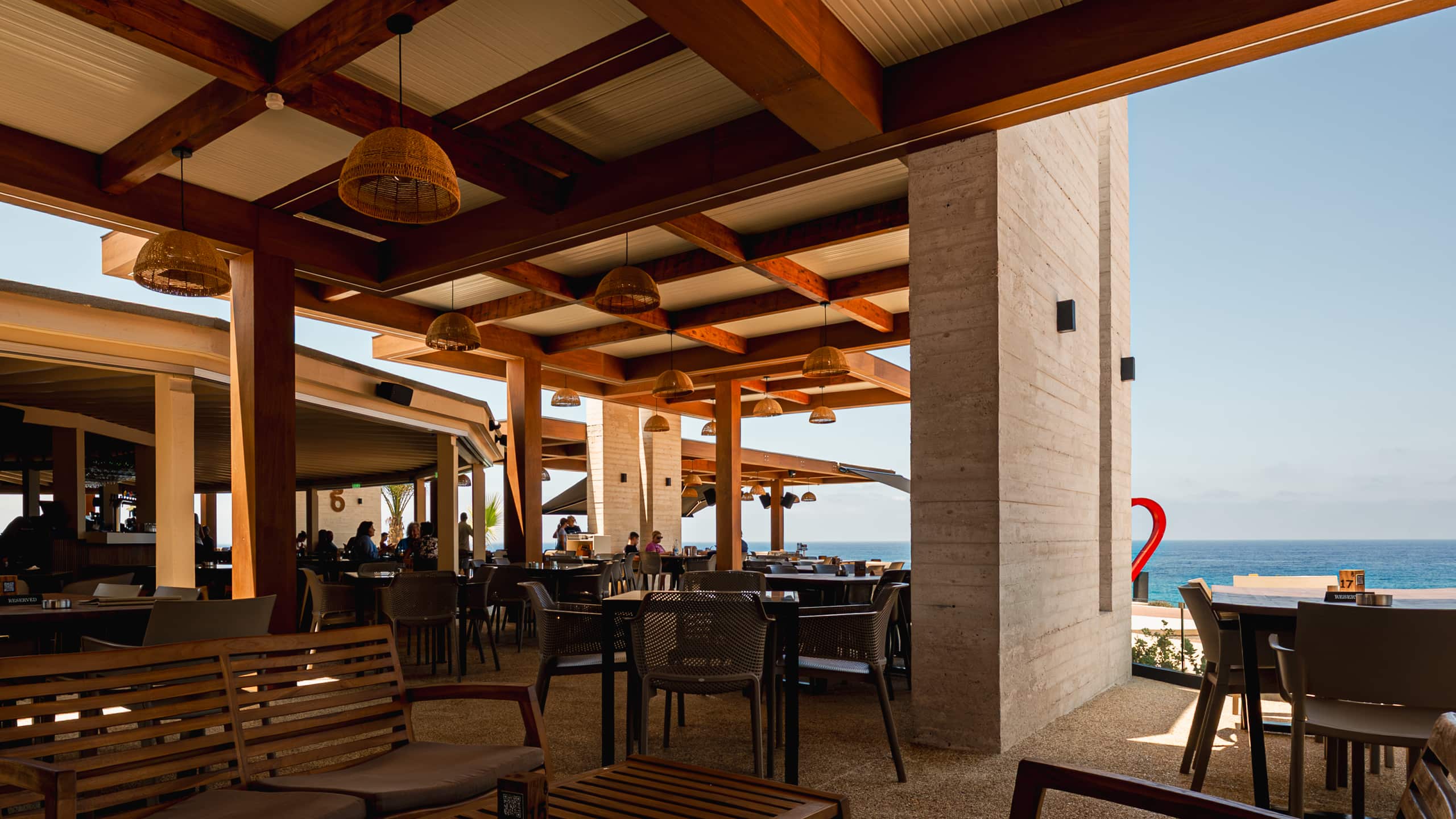 Under the Baracas pergola looking out to sea - exposed timber beams, rattan pendant lamps, laid tables and the red heart sculpture at the right edge.